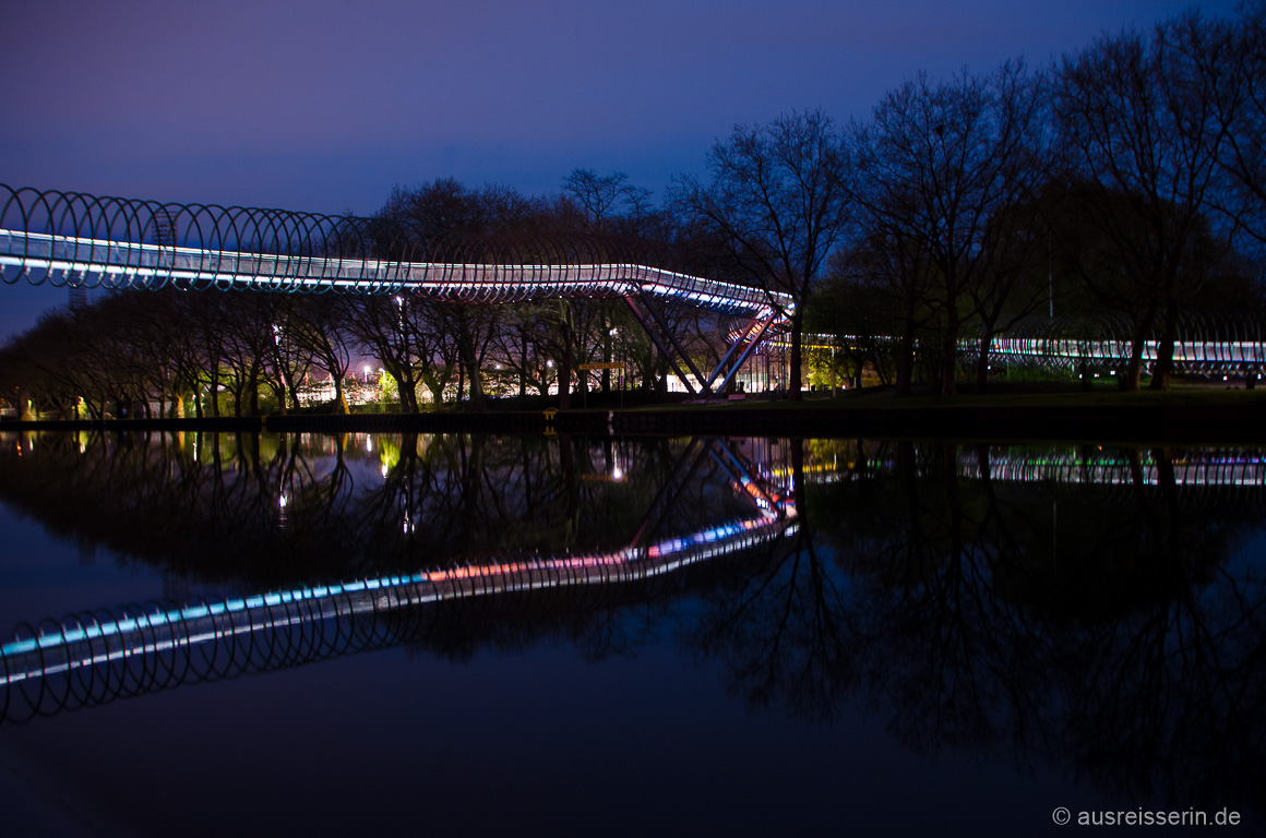 Die beleuchtete Spiralbrücke spiegelt sich im Wasser.
